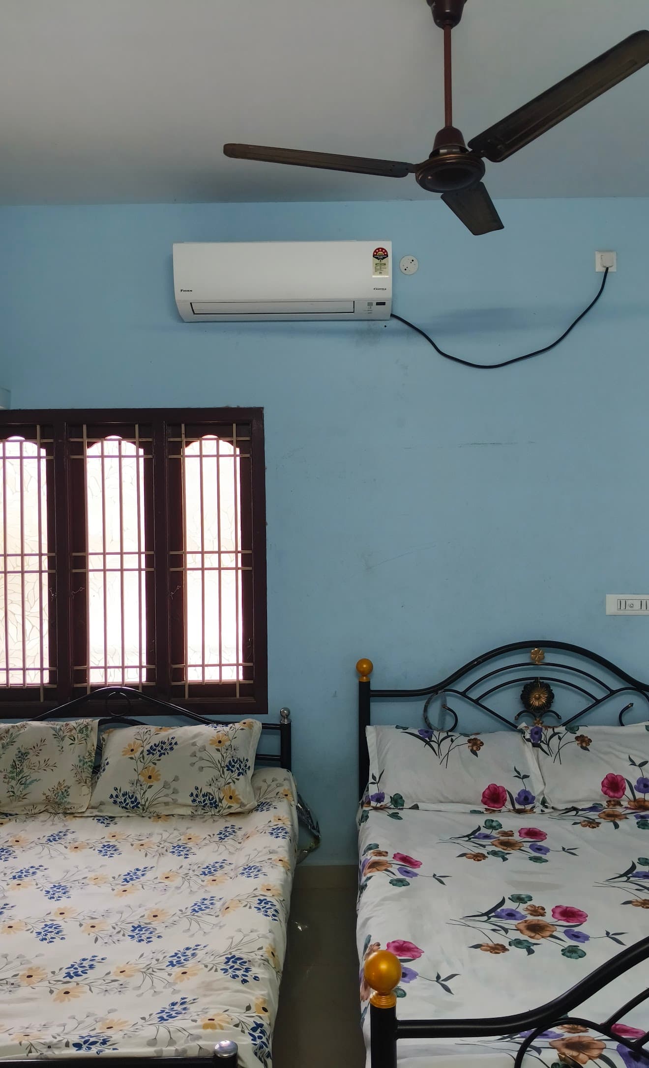 Blue bedroom featuring two floral-patterned beds, a wall-mounted air conditioner, and a ceiling fan.
